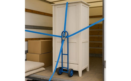 Armoire en bois clair fixée sur un diable bleu à l’intérieur d’un camion de déménagement, maintenue par des sangles à cliquet bleues, avec cartons et couvertures pliées en arrière-plan.